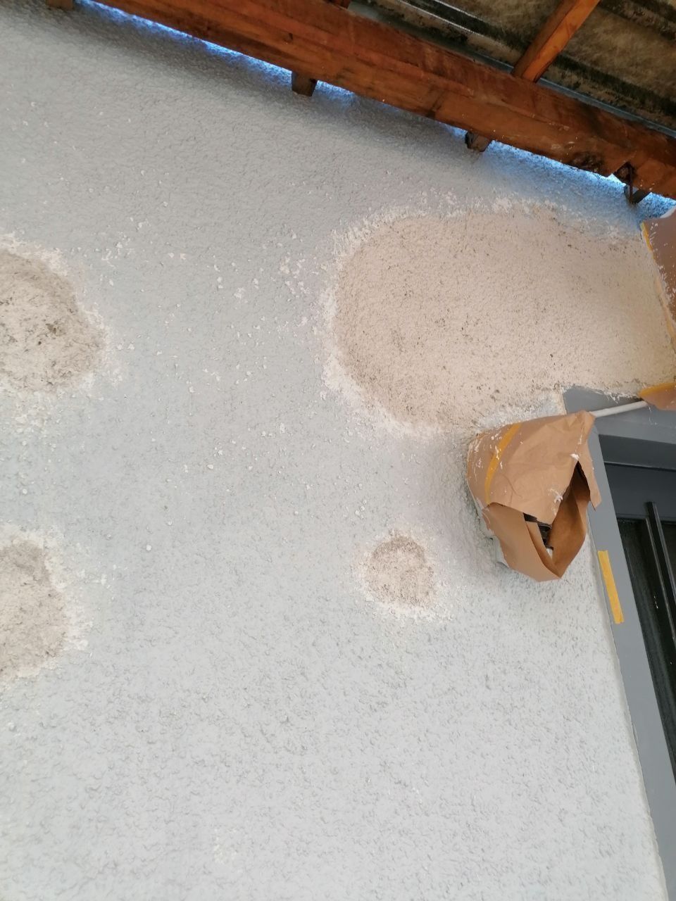 Hellblaue Außenwand mit mehreren Stellen freiliegenden Stucks. Eine braune Papiertüte hängt neben einer Tür.