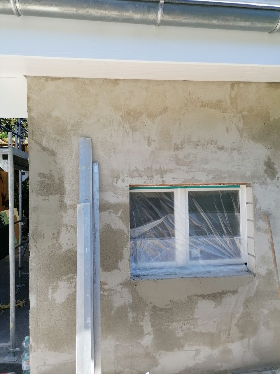 Außenwand im Bau mit einem mit Plastik abgedeckten Fenster. Baumaterialien stehen in der Nähe der Wand.
