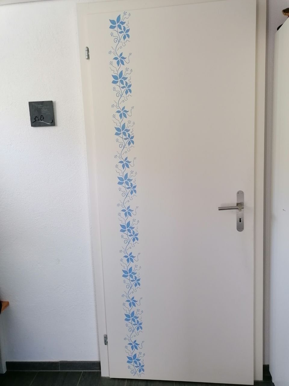 Weiße Tür mit blauem Blumenmuster auf der linken Seite. Auf der rechten Seite ist ein Metallgriff sichtbar.
