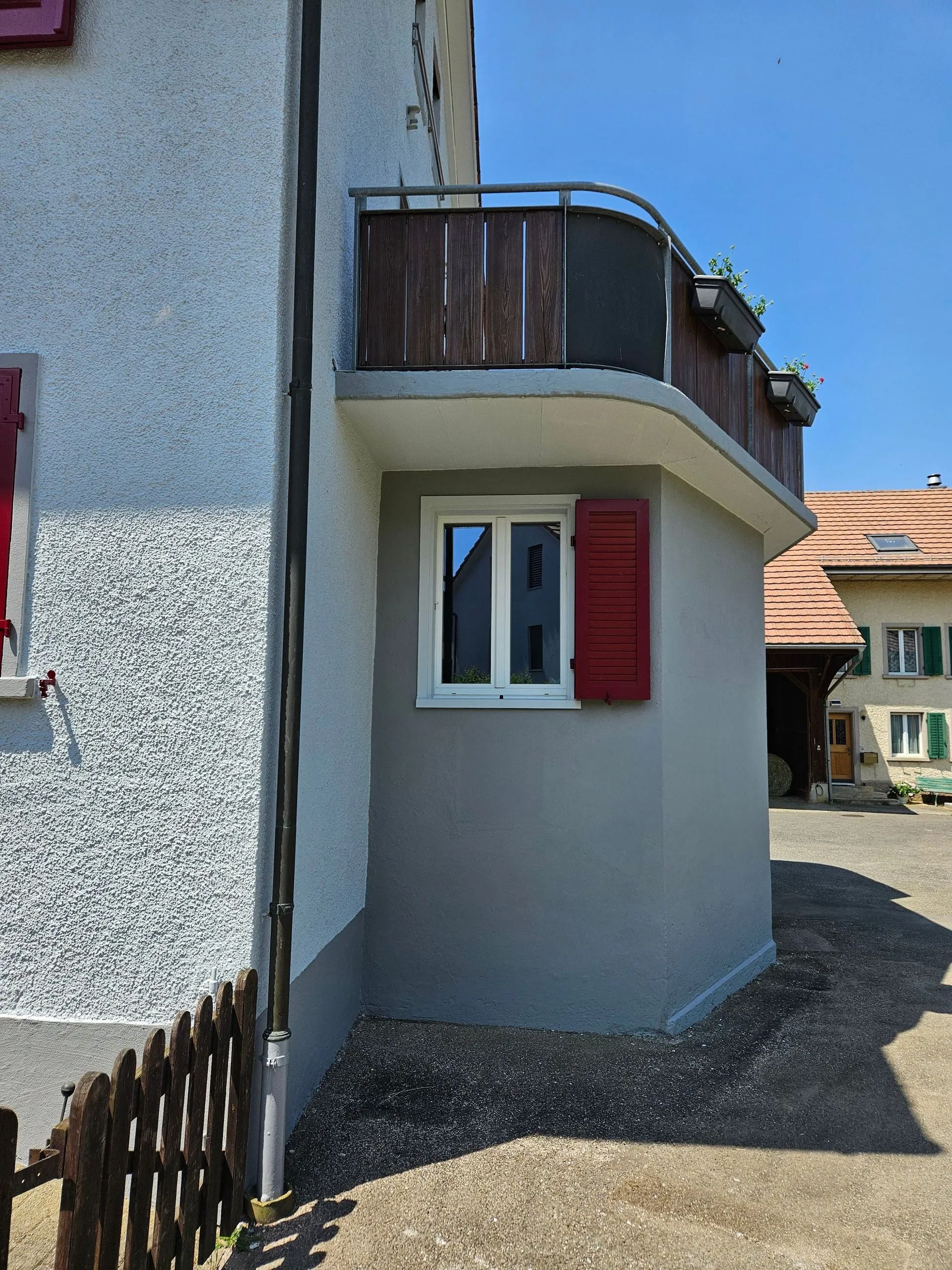 Gebäudeaußenseite mit kleinem Balkon, Fenster mit roten Fensterläden und einem Holzzaun.