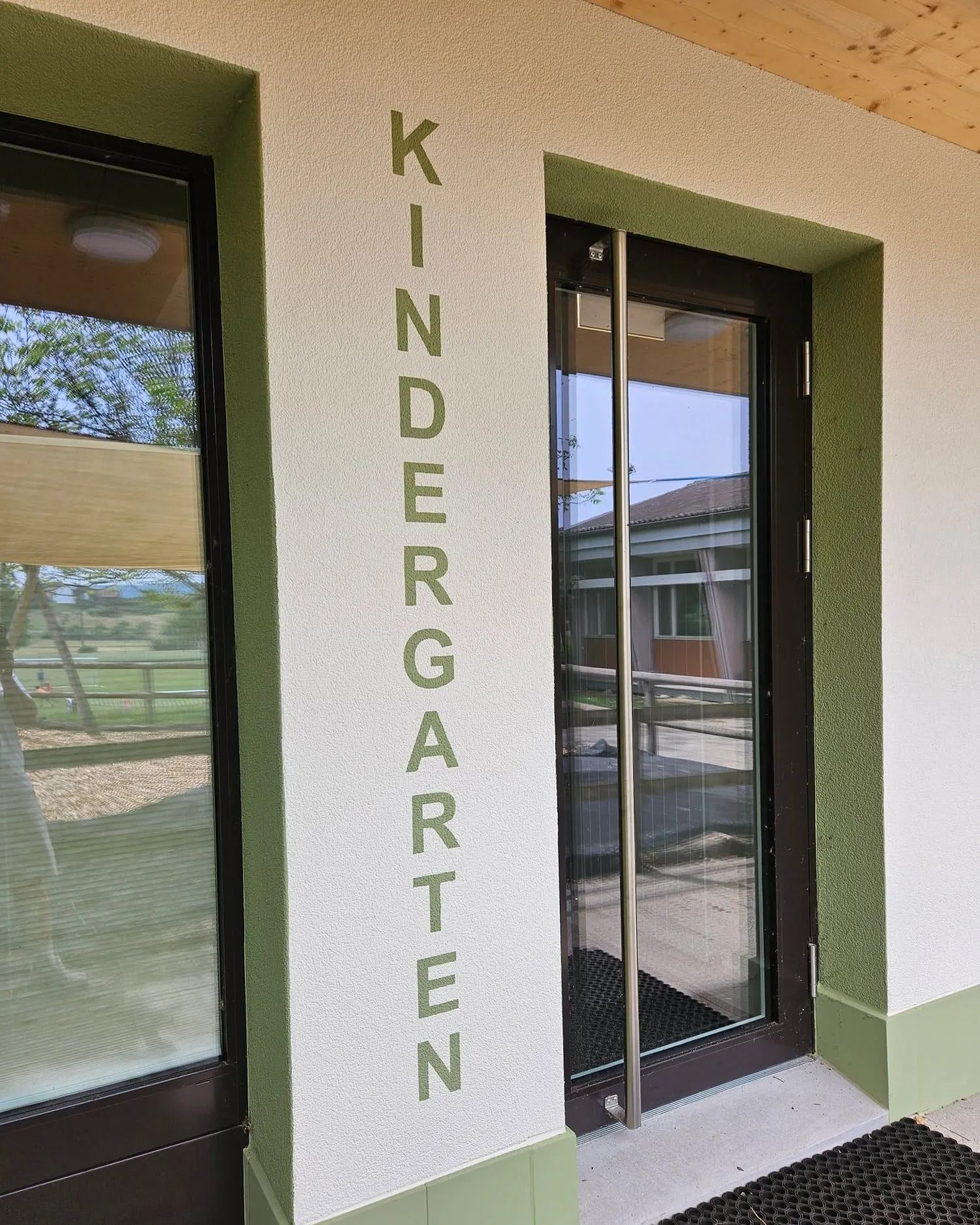 Eingang zu einem Kindergarten mit dunkelbrauner Tür, Glasfenstern und dem Wort „KINDERGARTEN“ in Grün.