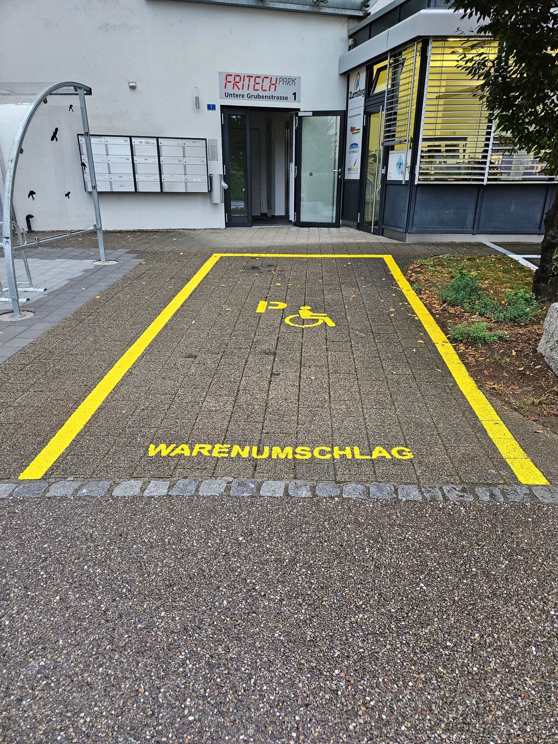 Gelb markierter Parkplatz mit Rollstuhlsymbol und der Aufschrift „WARENUMSCHLAG“ auf einer Schotterfläche vor einem Gebäude.