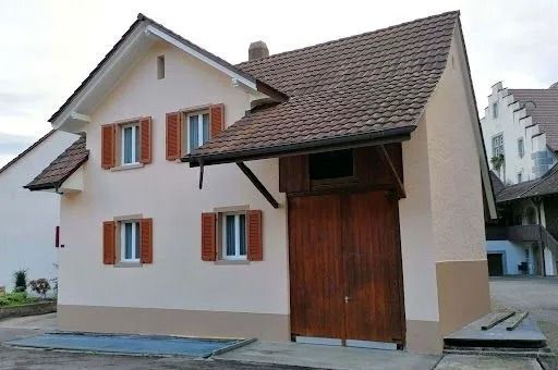 Hellbraunes Gebäude mit braunen Fensterläden und einer großen Holztür unter einem Ziegeldach.