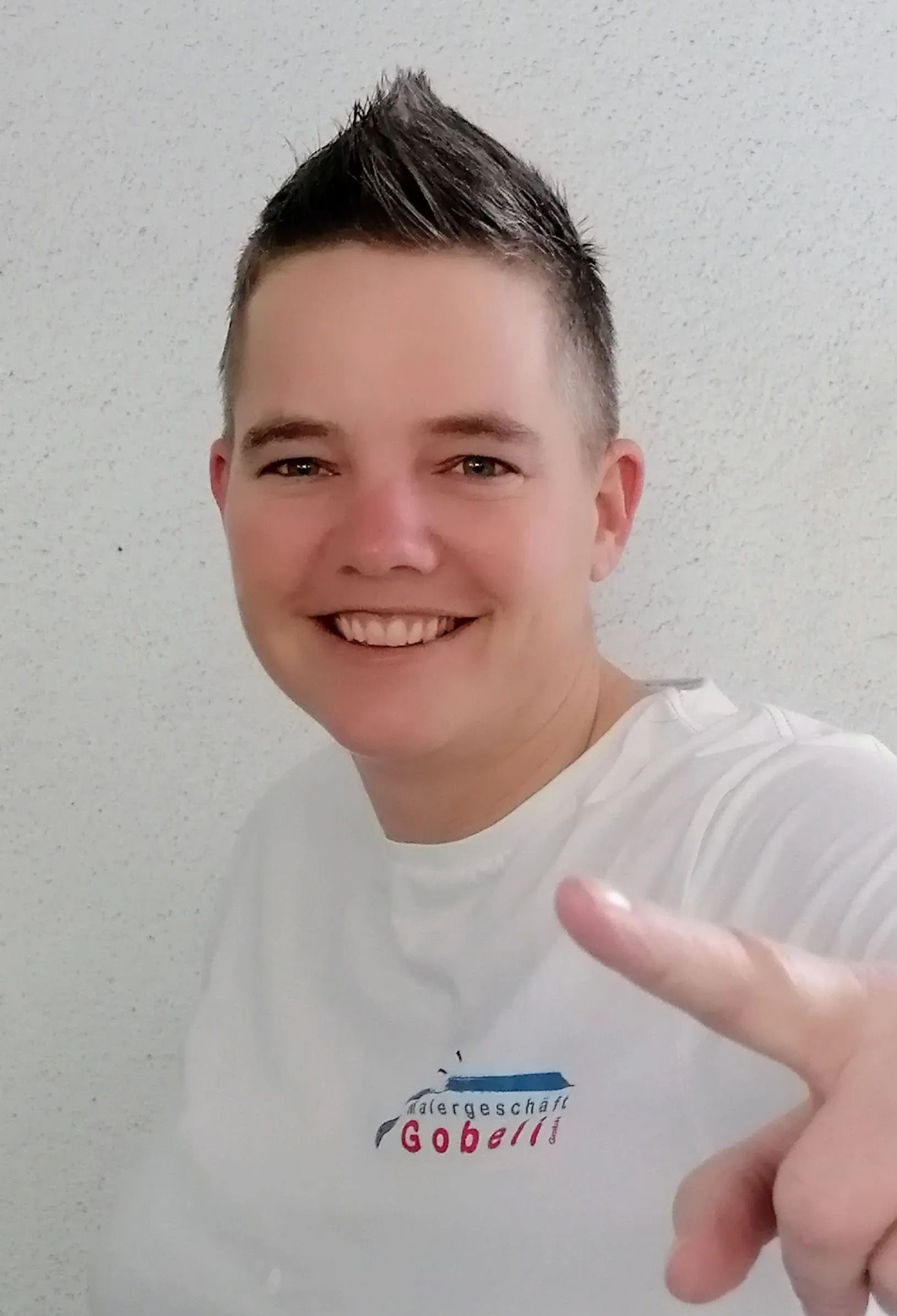 Person mit stacheligem Haarschnitt lächelt und zeigt mit dem Finger. Trägt ein weißes T-Shirt vor einer weißen Wand.