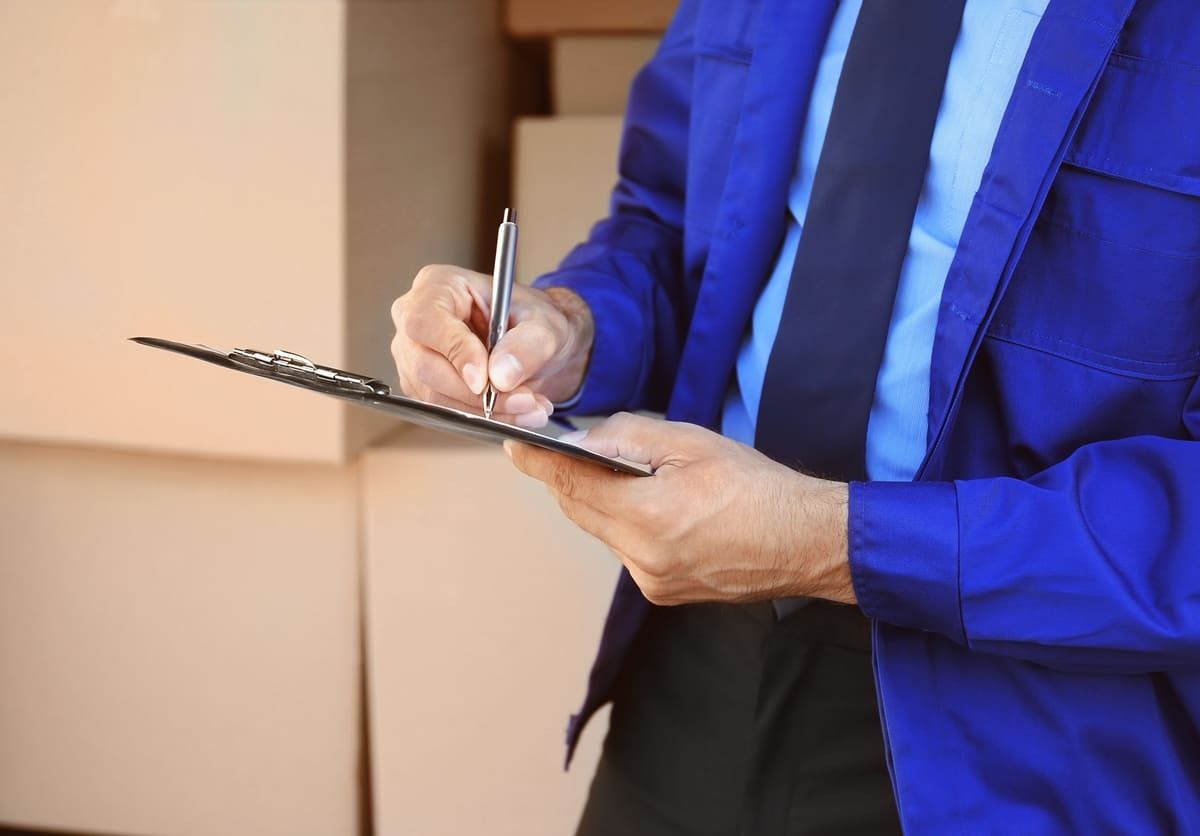 Un hombre con uniforme azul firma un portapapeles delante de unas cajas de cartón.