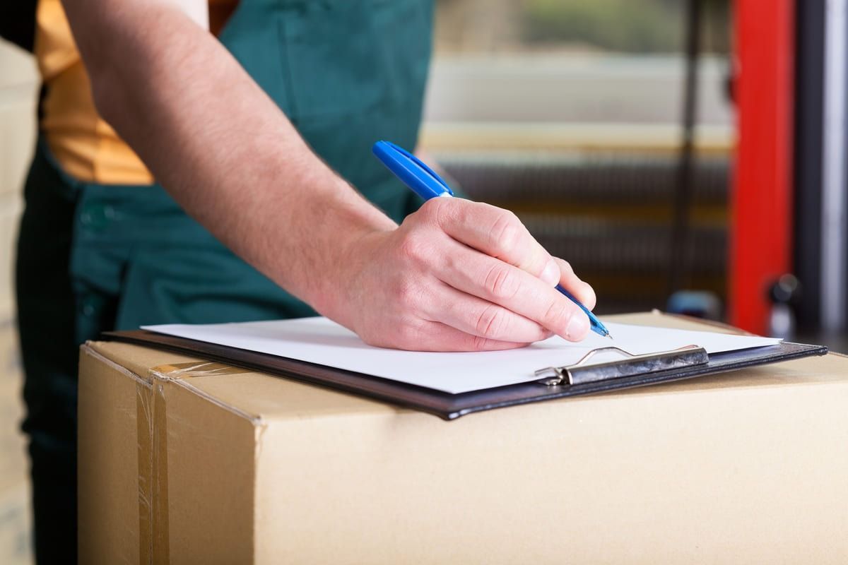 Persona con mono verde firmando en un portapapeles encima de una caja de cartón.