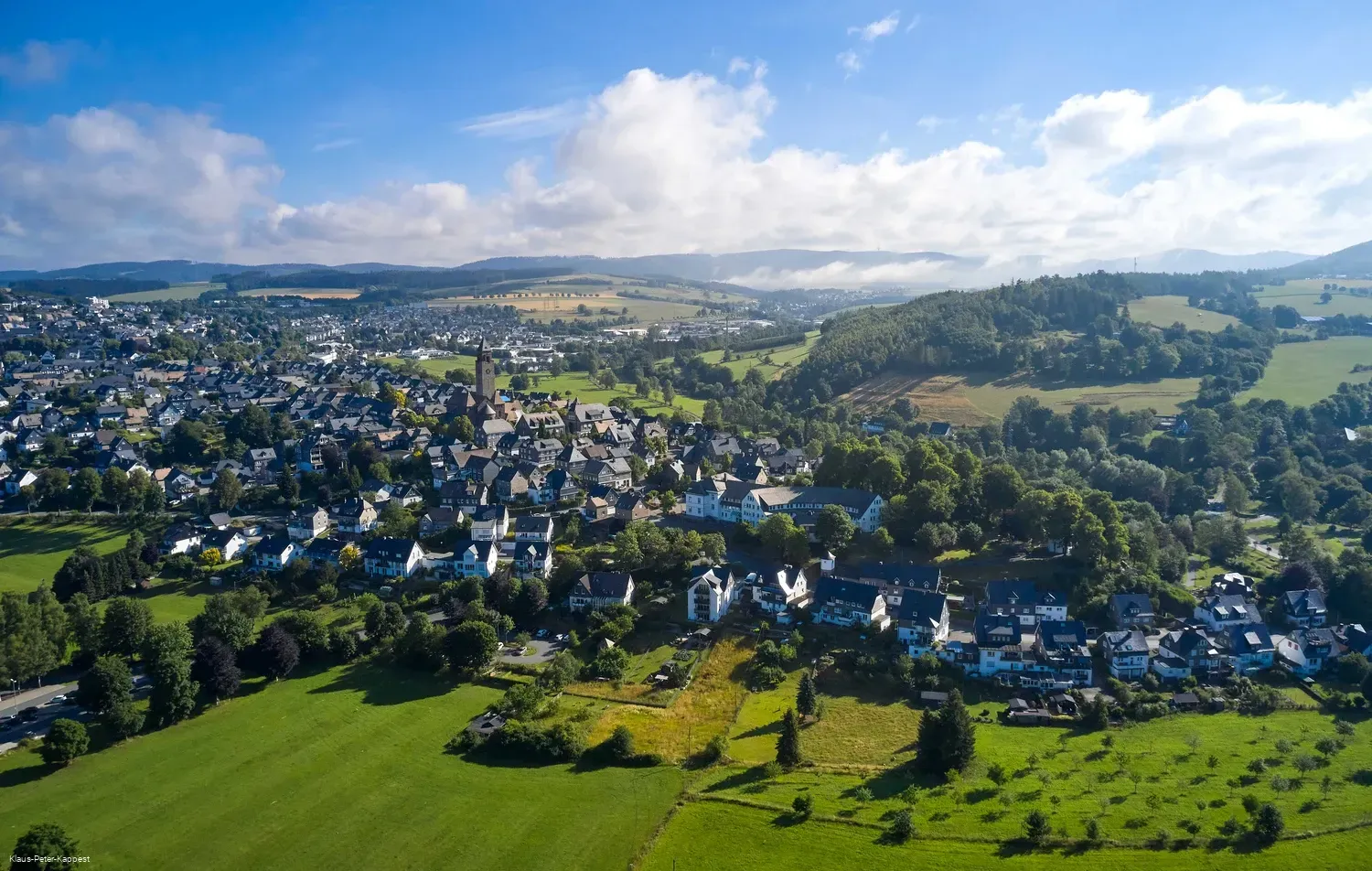 Luftaufnahme einer Stadt, eingebettet in ein Tal mit grünen Feldern und sanften Hügeln unter blauem Himmel.