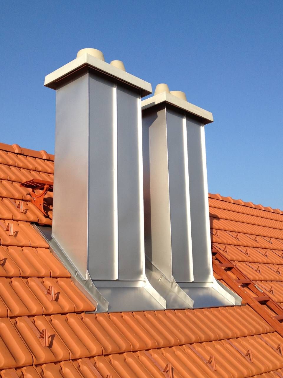 Zwei glänzende Metallschornsteine ​​auf einem roten Ziegeldach vor klarem, blauem Himmel.