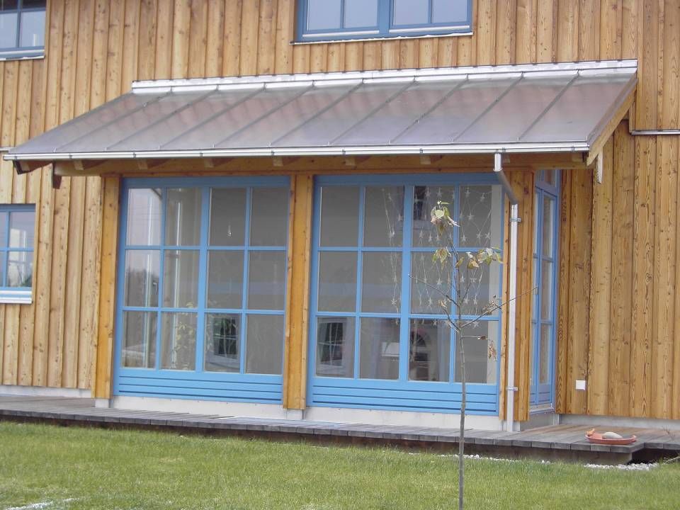Eine mit blauem Rahmen versehene Glasveranda mit transparentem Dach ist an ein Holzgebäude auf einem Rasen angebaut.