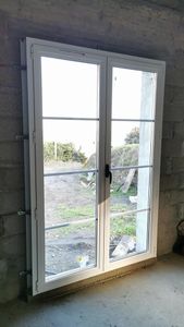 Portes-fenêtres blanches à double vitrage avec grilles installées dans une pièce en béton brut donnant sur une cour gazonnée.
