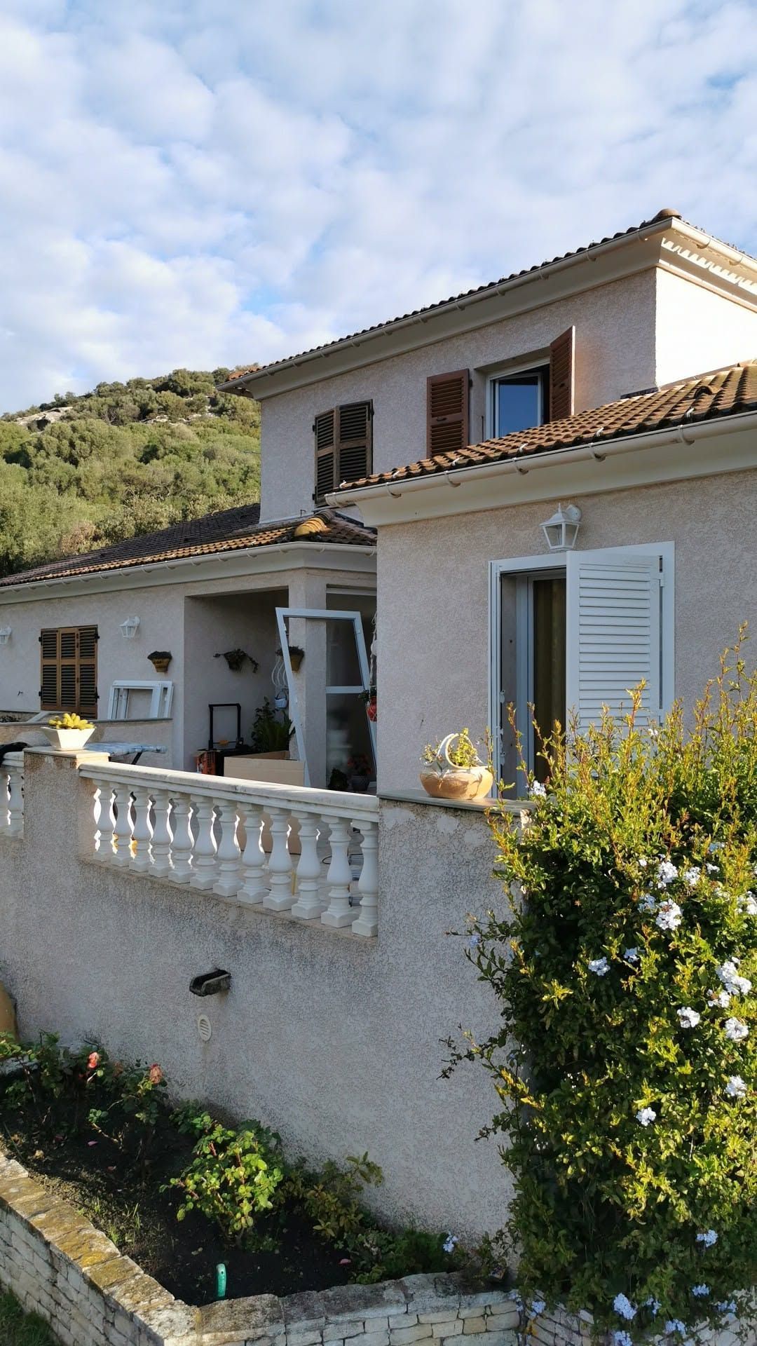 Une maison blanche à deux étages avec une façade arrondie, des fenêtres sombres, une balustrade de balcon couleur pierre et des pins.