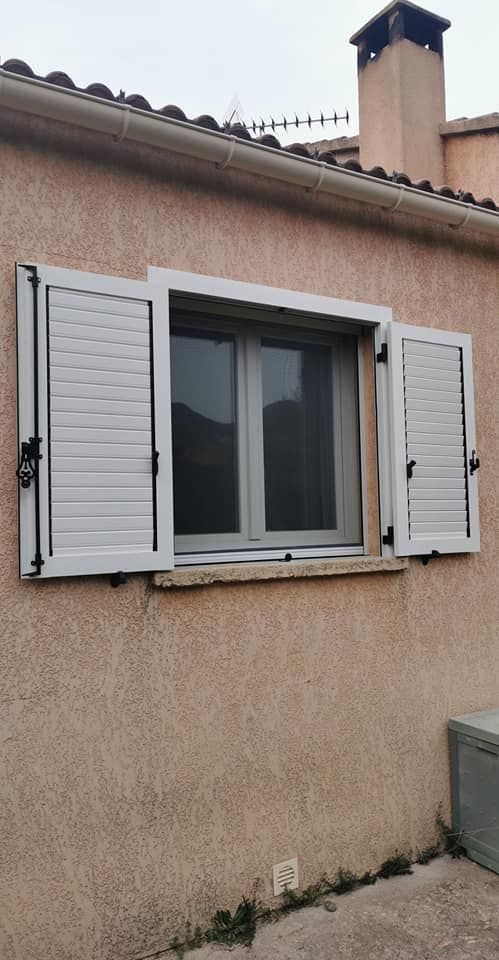 Une fenêtre carrée aux cadres gris et aux volets blancs s'ouvre sur un mur extérieur texturé, de couleur brun clair.