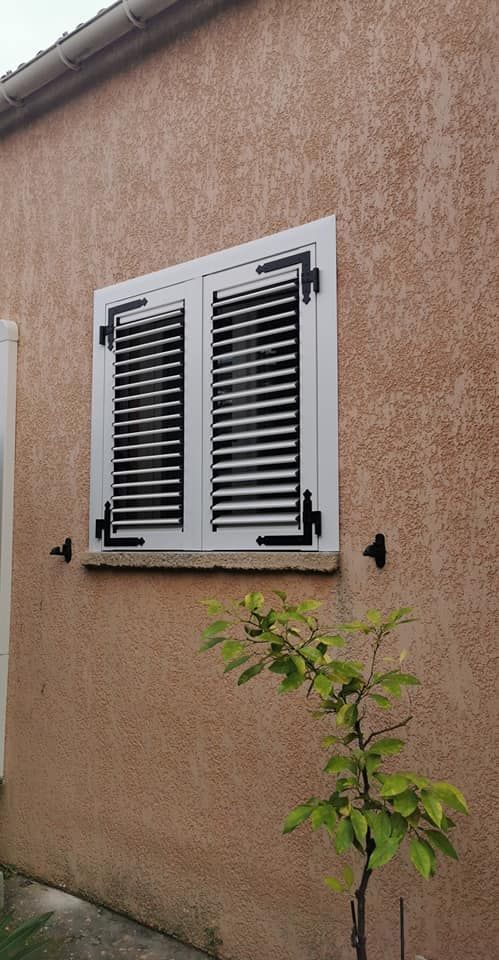 Une fenêtre blanche à persiennes noires sur un mur extérieur texturé brun clair, à côté d'une petite plante verte.