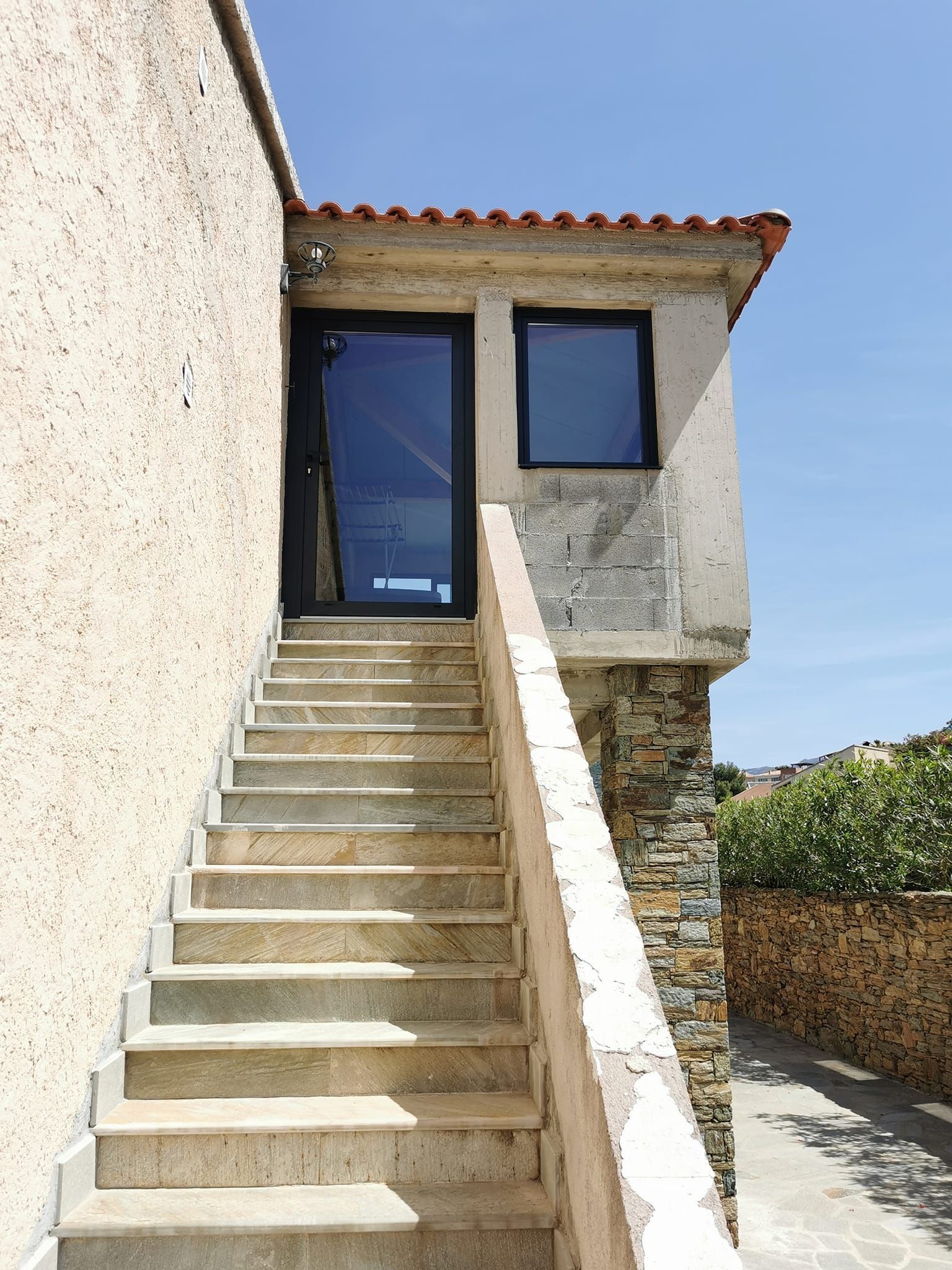 Un escalier en pierre menant à l'entrée d'un petit bâtiment avec une porte et une fenêtre sombres se détachant sur un ciel bleu clair.