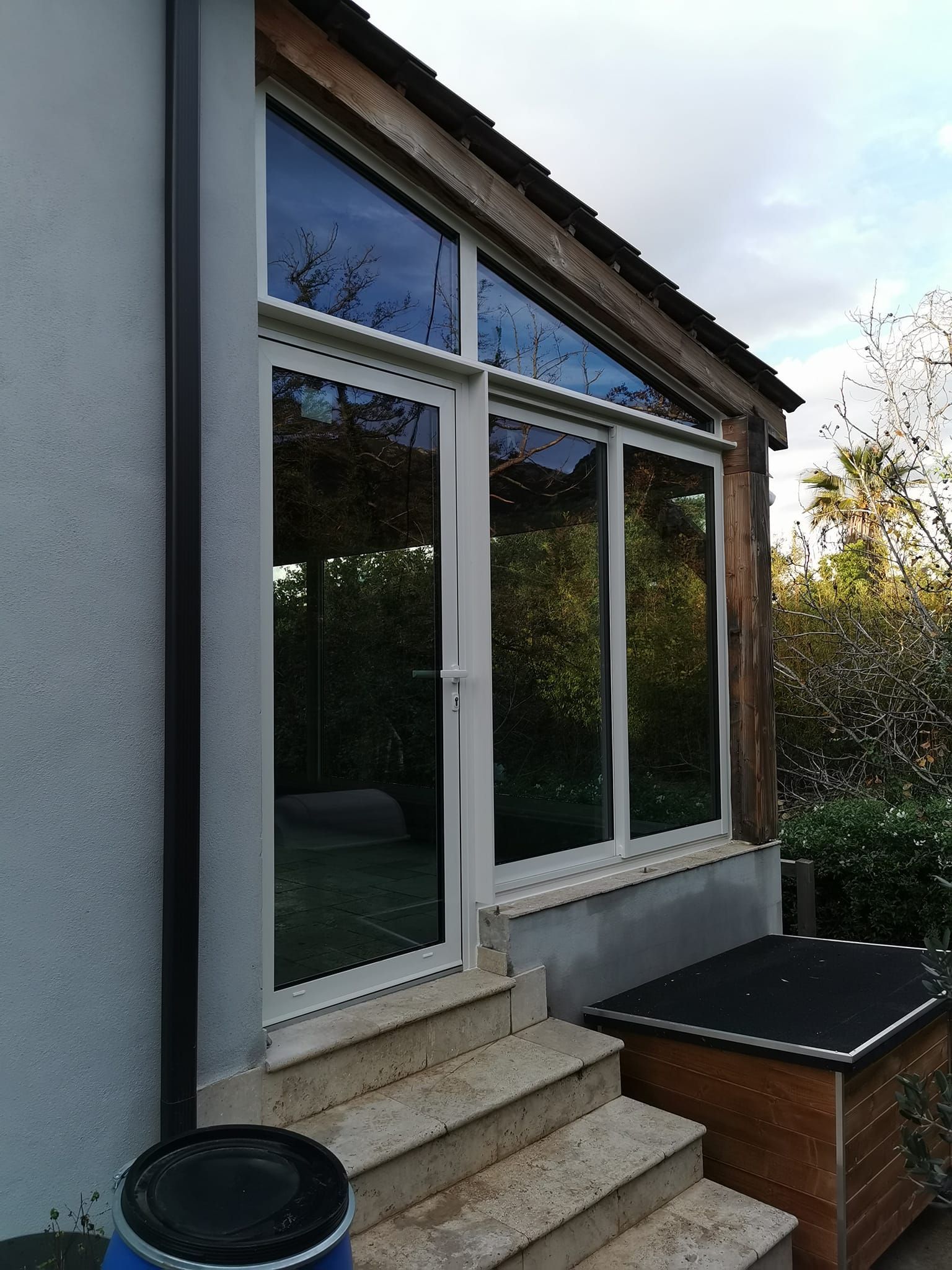 Une porte vitrée à cadre blanc et une fenêtre latérale sont placées sur un escalier en pierre, contre un mur gris clair et une poutre en bois.