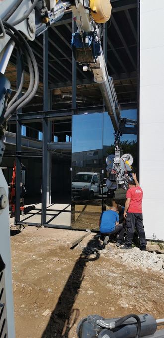 Deux ouvriers du bâtiment guident un grand panneau de verre, soulevé par un système de vide, jusqu'à sa mise en place sur la façade d'un bâtiment à ossature métallique.