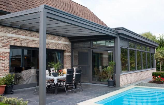 Une terrasse avec une pergola gris foncé et une véranda vitrée attenante à une maison en briques, à côté d'une piscine extérieure.