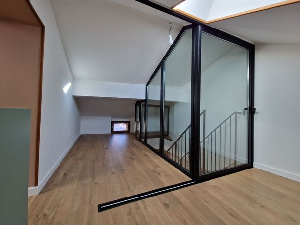 Un espace intérieur moderne avec un sol imitation bois, des murs blancs et une cloison vitrée à cadre noir qui renferme un escalier.