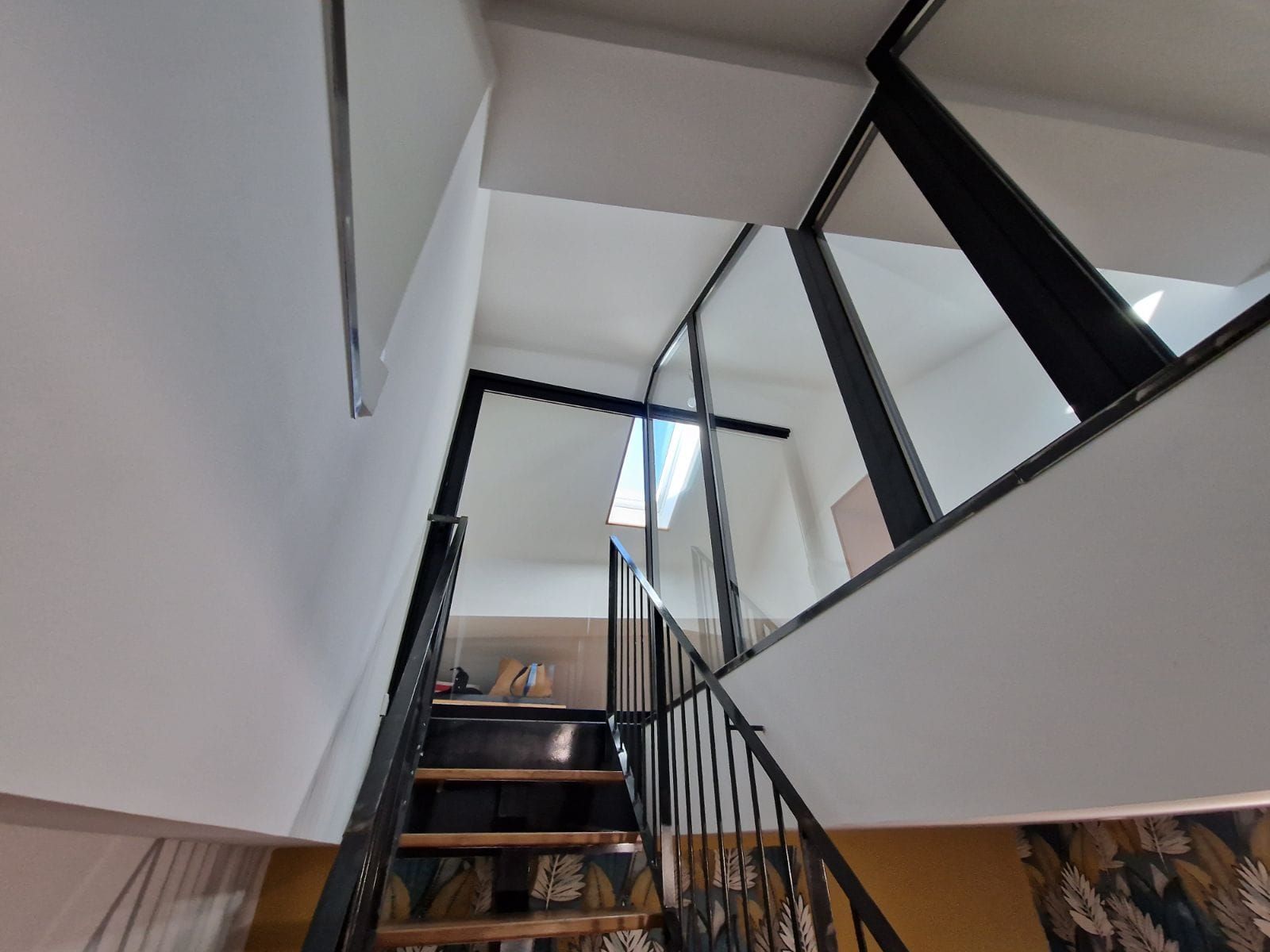 Un escalier aux marches en bois et à la rampe en métal noir mène vers une lucarne dans un intérieur aux murs blancs.