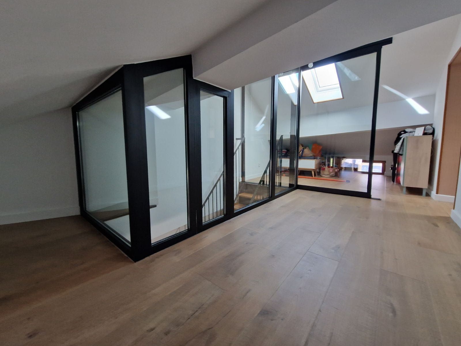 Une pièce mansardée moderne avec parquet clair et parois vitrées à cadre noir abritant un escalier menant à un niveau inférieur.