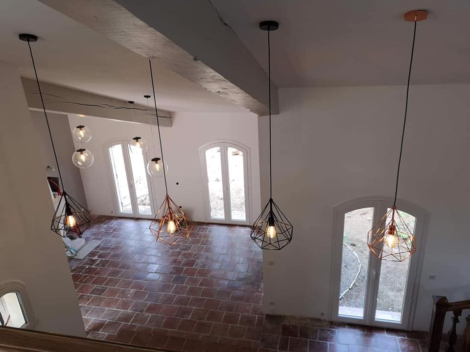 Trois suspensions lumineuses géométriques modernes éclairent une pièce baignée de soleil, avec un sol en terre cuite et des portes-fenêtres cintrées.
