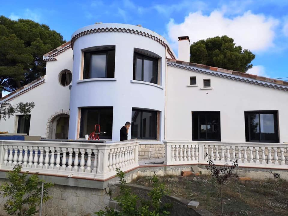 Une maison blanche à deux étages avec une façade arrondie, des fenêtres sombres, une balustrade de balcon couleur pierre et des pins.