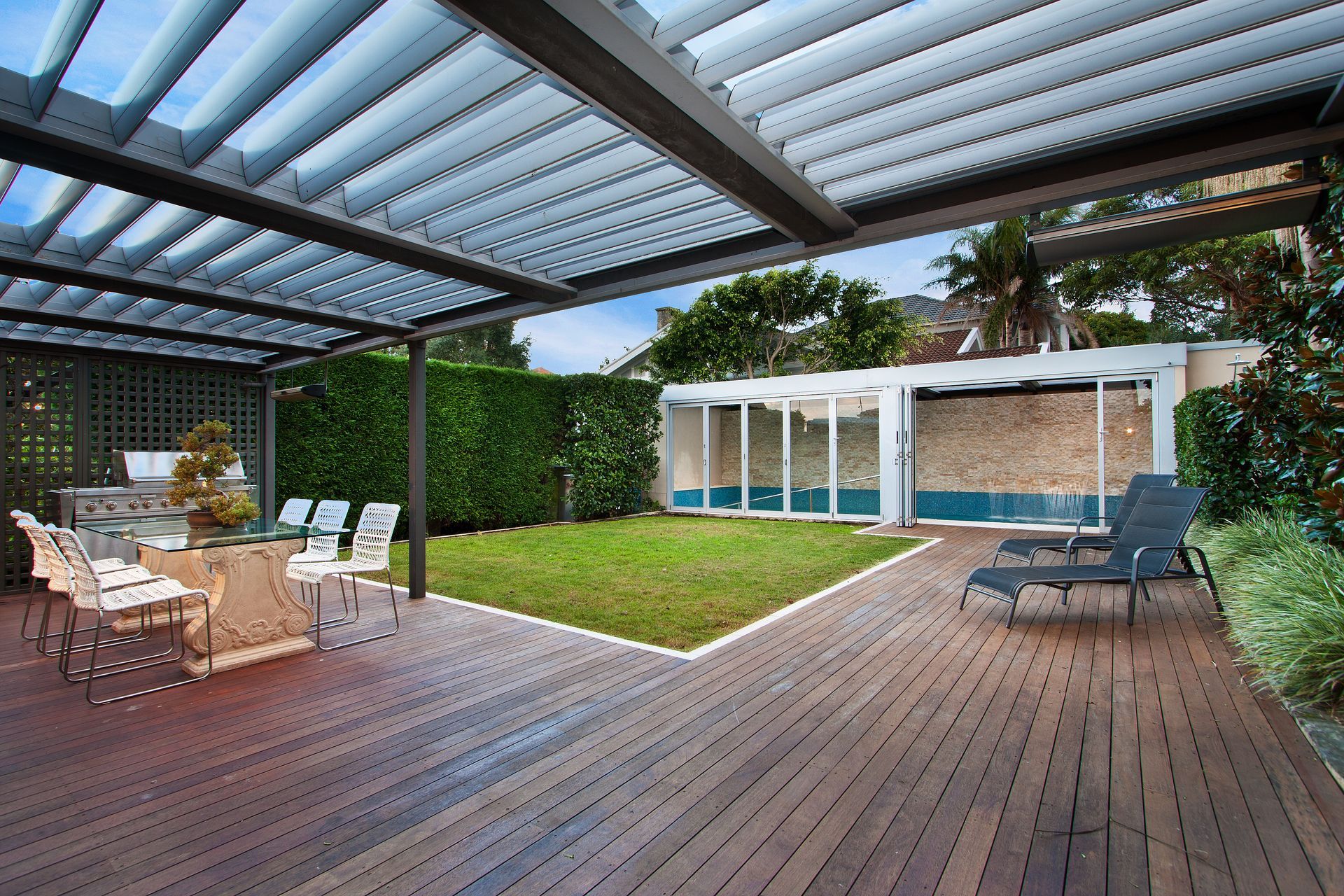 Terrasse extérieure avec terrasse en bois, pergola, ensemble de salle à manger et pelouse menant à un espace piscine avec portes vitrées.