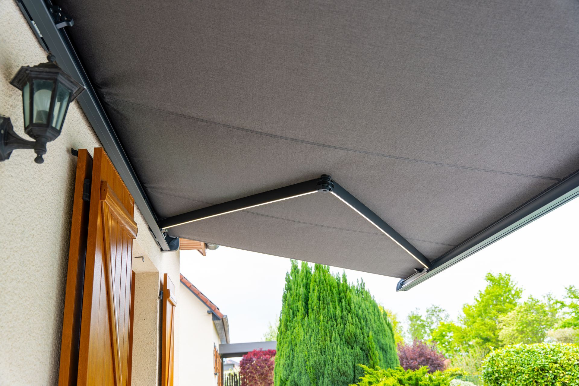 Un auvent de terrasse rétractable couleur anthracite s'étendait au-dessus de la façade d'un bâtiment, avec des volets en bois et une applique murale.