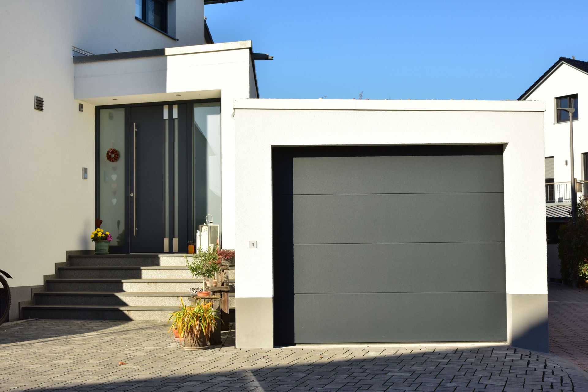 L'extérieur d'une maison moderne blanche, avec des marches en pierre menant à une porte d'entrée gris foncé, jouxtant une porte de garage de même couleur.
