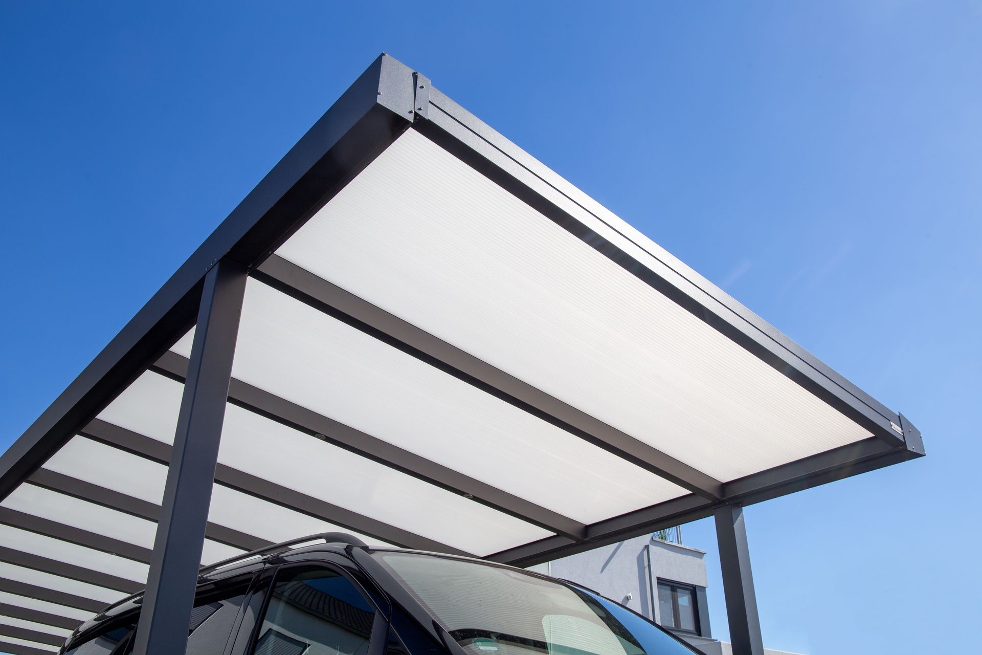 Un abri de voiture à structure sombre et à panneaux de toit translucides abrite un véhicule sombre sous un ciel bleu clair.