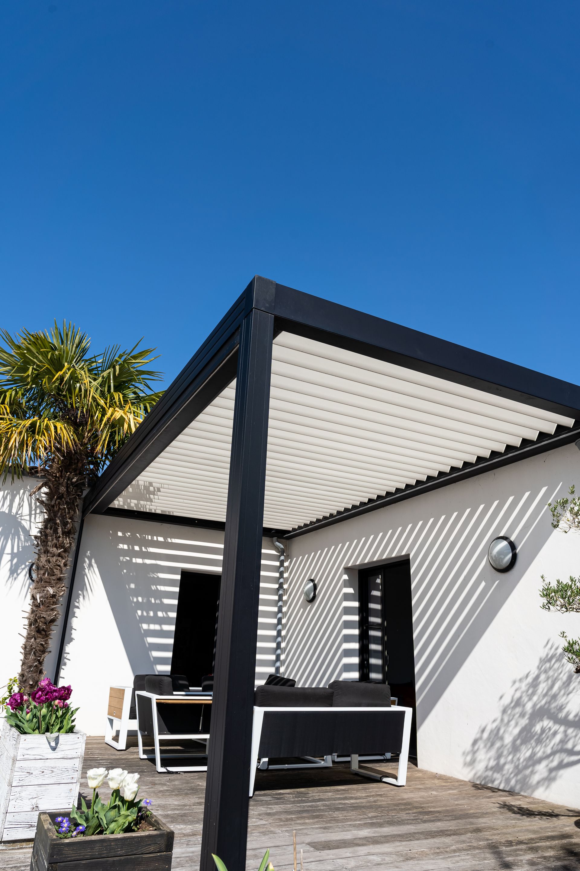 Une pergola moderne à structure noire et à persiennes blanches ombrage une terrasse adossée à un mur blanc sous un ciel bleu azur.