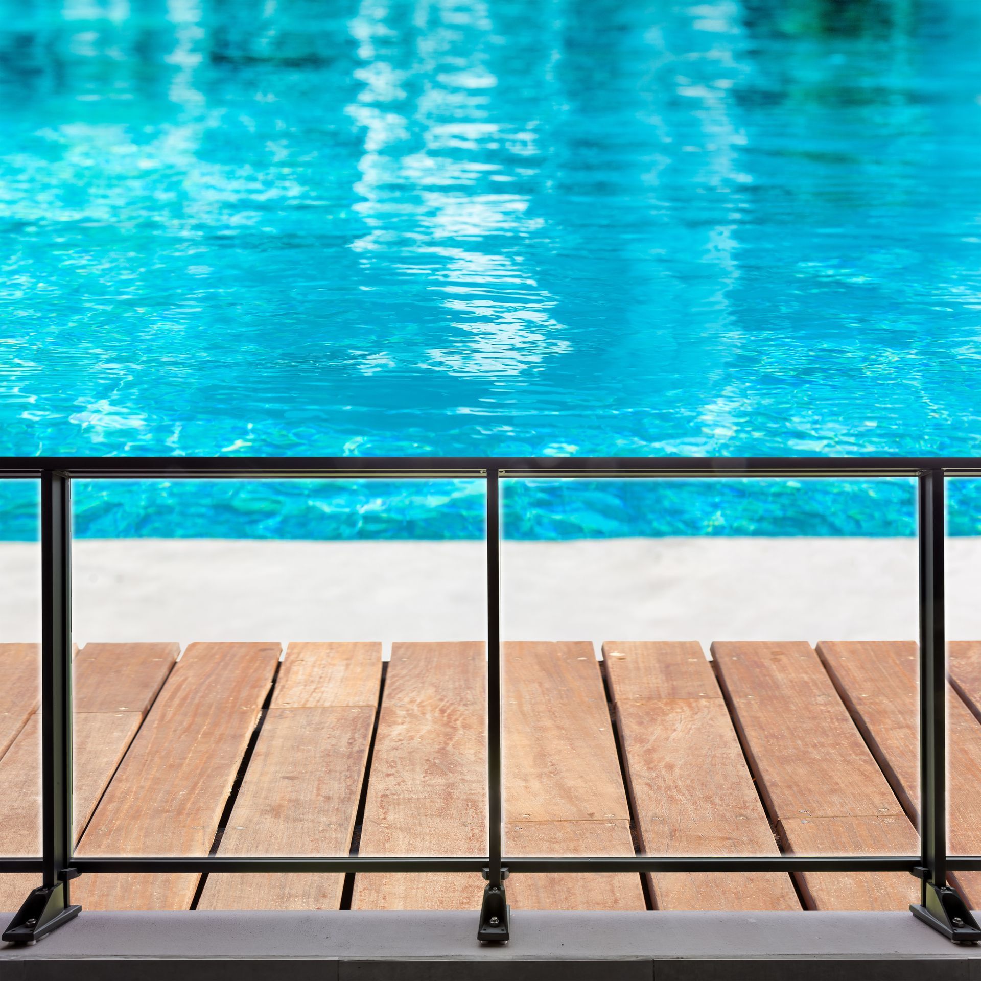 Une clôture en métal sombre se dresse devant une terrasse en bois qui surplombe une piscine d'un bleu éclatant.