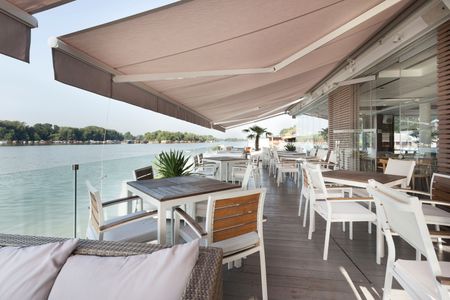 Terrasse extérieure de restaurant au bord de l'eau avec chaises blanches, tables et un grand auvent beige donnant sur une rivière calme.