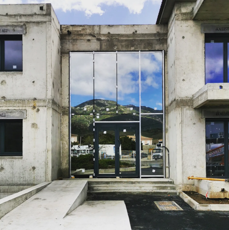 Entrée d'un bâtiment en béton avec un panneau central en verre reflétant une montagne et le ciel, comportant des portes doubles et une rampe.
