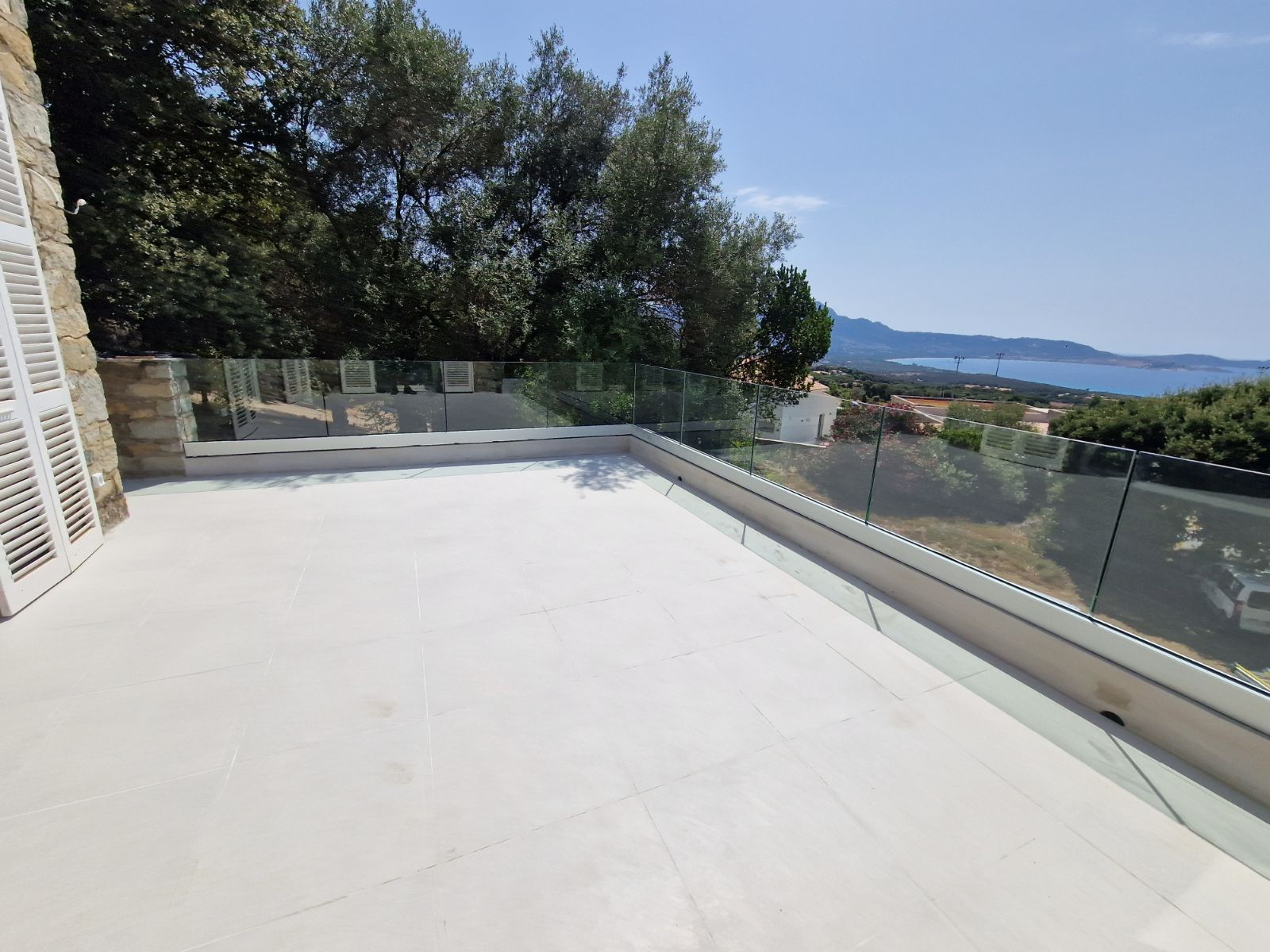 Un spacieux balcon en pierre blanche avec une balustrade en verre sans cadre donnant sur un paysage côtier pittoresque et une végétation luxuriante.