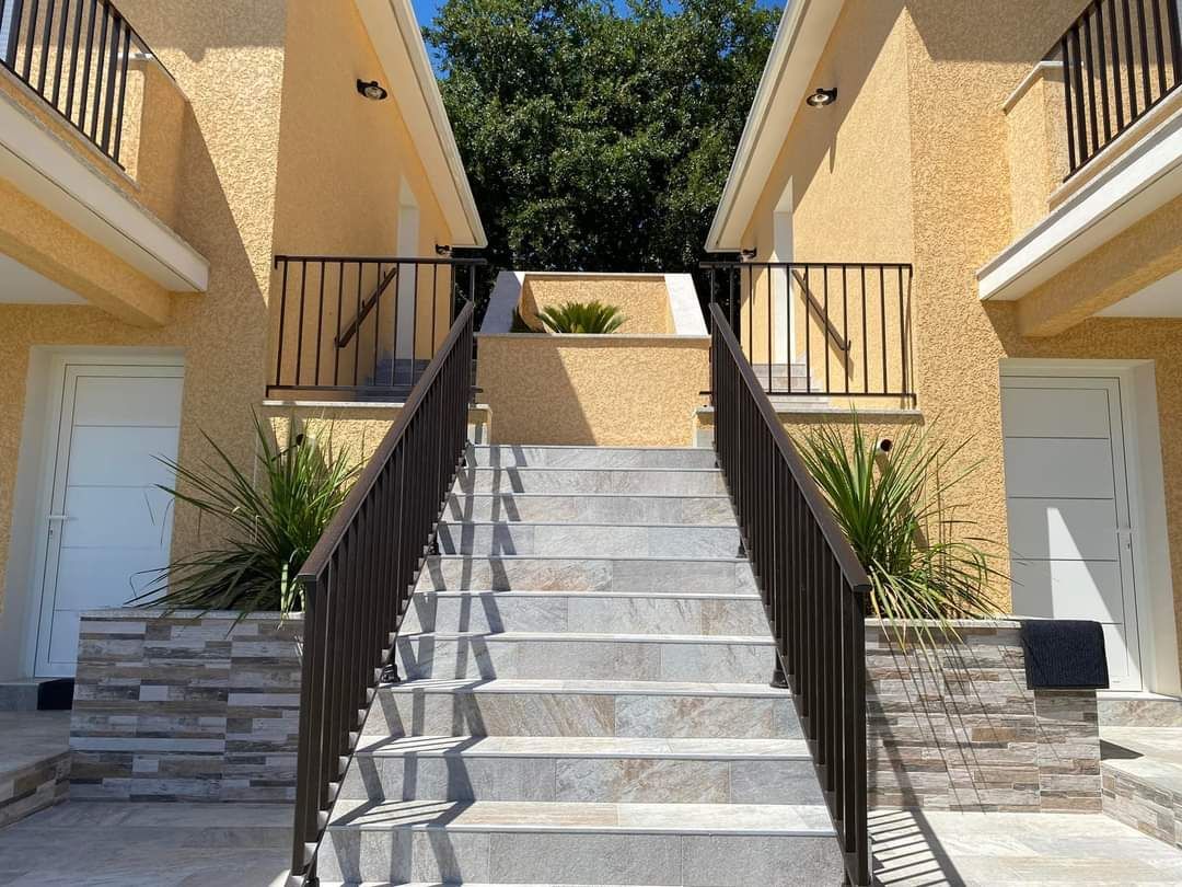 Un large escalier en pierre avec des rampes en métal noir mène entre deux bâtiments beiges aux portes blanches et ornés de petites plantes.