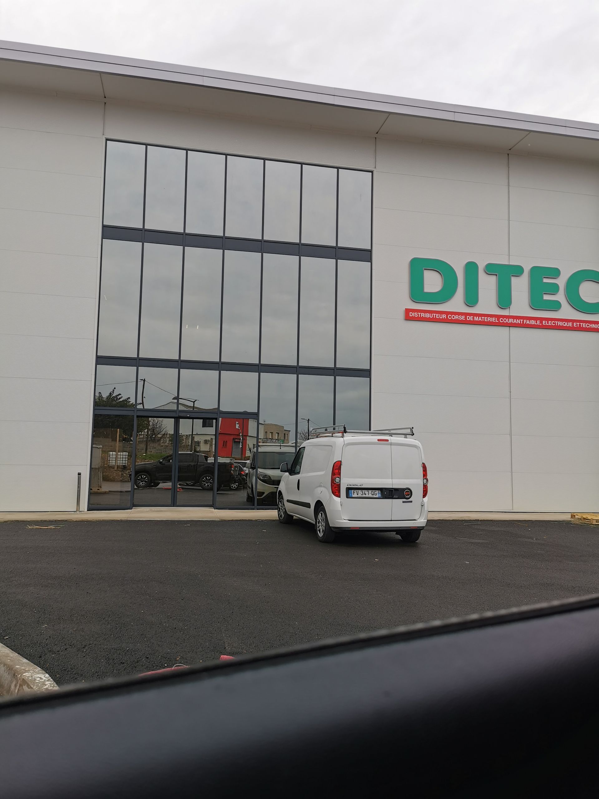 Un bâtiment commercial blanc de DITEC avec une haute façade vitrée et une camionnette blanche garée sur le parking en gravier devant.