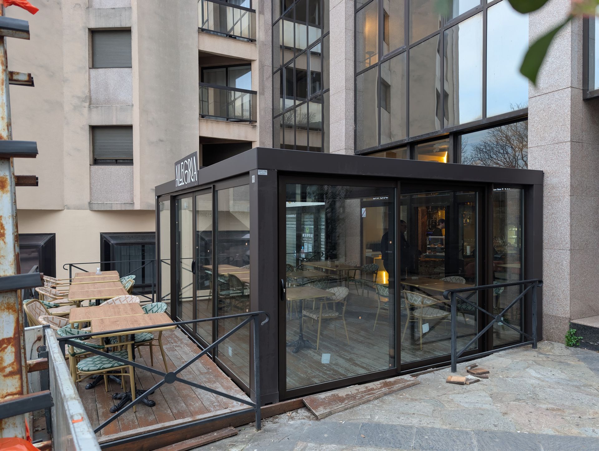 Un espace terrasse extérieure aménagé dans une structure moderne en verre à cadre sombre, jouxtant un immeuble de plusieurs étages.