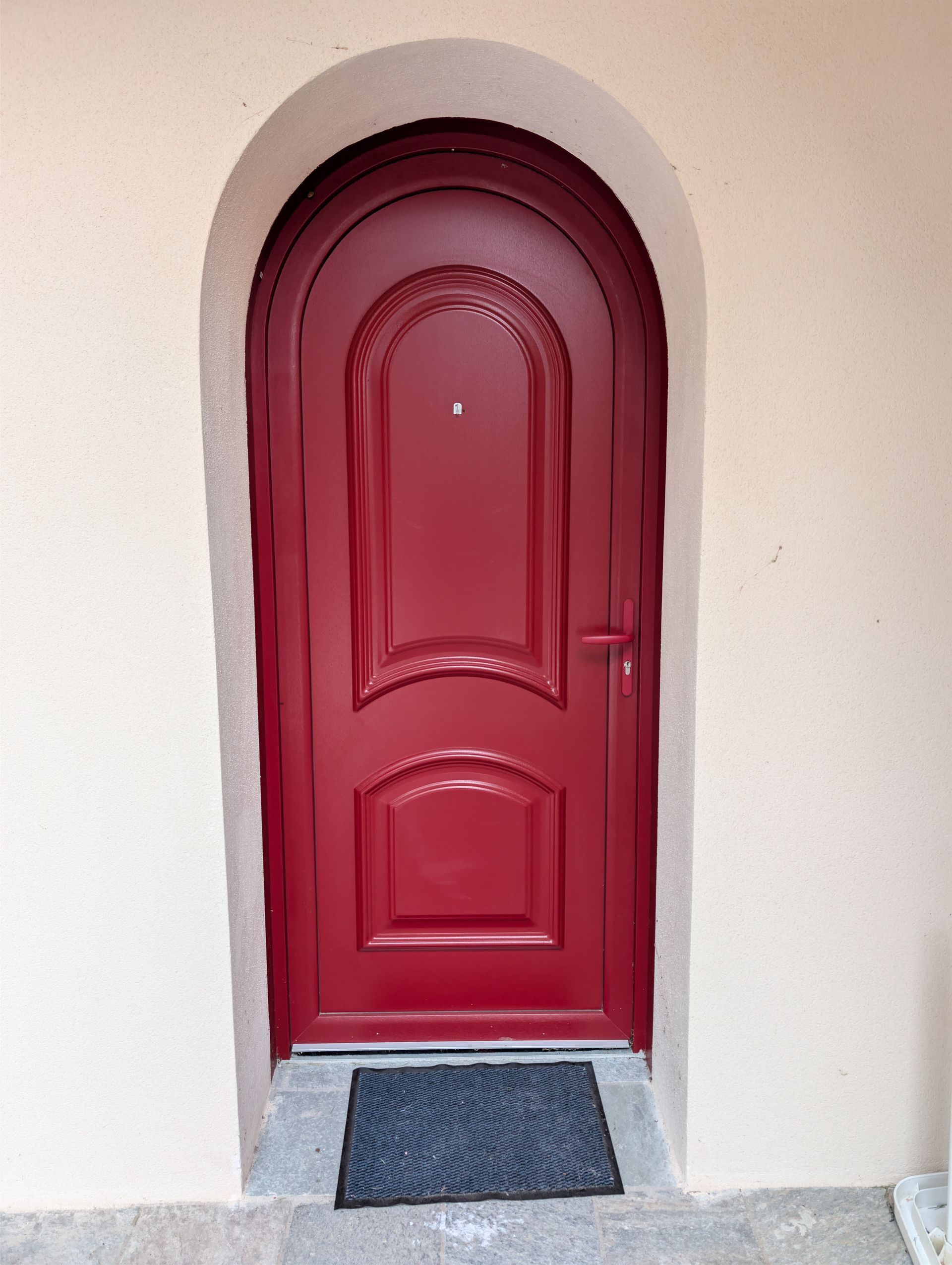 Une porte d'entrée rouge en arc de cercle, centrée dans un mur beige, avec un paillasson gris au sol devant.