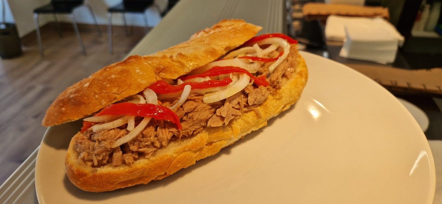 Sándwich con carne en rodajas y hierbas sobre una tabla de madera, acompañado de un pequeño plato de pimientos verdes.