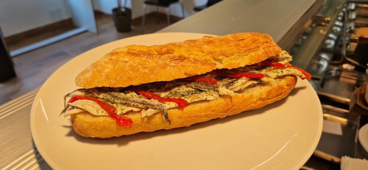 Dos bocadillos de baguette sobre una tabla de madera con un pequeño cuenco de salsa junto a una ventana.