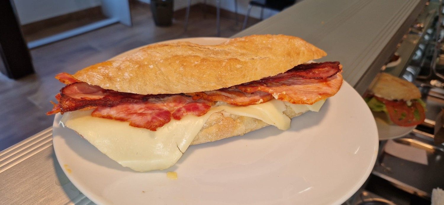 Sándwich con carne en rodajas y hierbas sobre una tabla de madera, acompañado de un pequeño plato de pimientos verdes.