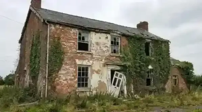 Casa de tijolos dilapidada, coberta de hera, janelas quebradas, porta entreaberta, em um campo tomado pelo mato.