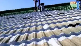 Uma pessoa utiliza um maçarico para instalar material de cobertura em um telhado plano. Um rolo de material é visível.