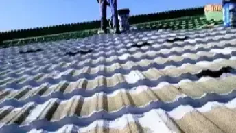 Uma pessoa utiliza um maçarico para instalar material de cobertura em um telhado plano. Um rolo de material é visível.