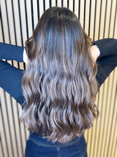 Una persona con cabello castaño largo y ondulado, vista de espaldas, con una blusa negra, apoyada contra una pared a rayas.