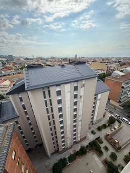 Vista aérea desde un ángulo elevado de un edificio de apartamentos alto, de color beige y con tejado de tejas oscuras, situado en una zona urbana densamente poblada.