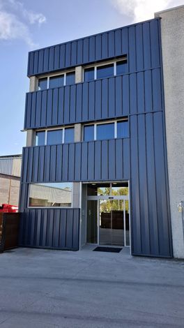 Fachada de un edificio moderno de tres plantas con paneles verticales de color azul oscuro y ventanas de cristal sobre una entrada en la planta baja.
