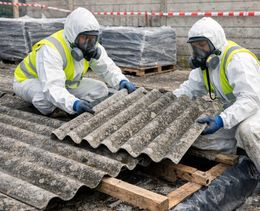 Dos trabajadores, equipados con trajes de protección completos y respiradores, retiran cuidadosamente paneles corrugados de una obra al aire libre.