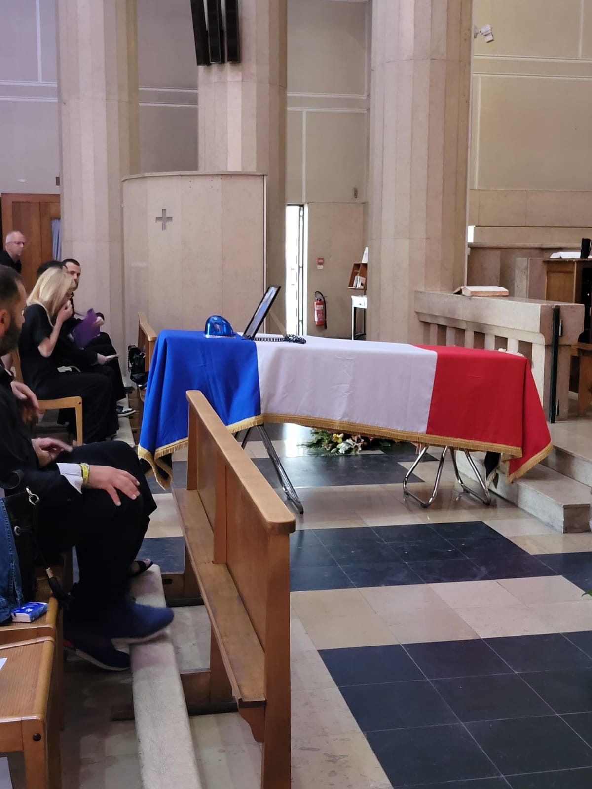 Cérémonie funéraire : cercueil recouvert du drapeau français ; personnes en deuil assises dans l’église.